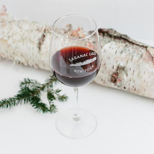 Wine glass with stem and SARANAC LAKE NEW YORK etched in white around mountains. Red beverage inside glass which stands in front of a birch log and pine branch.