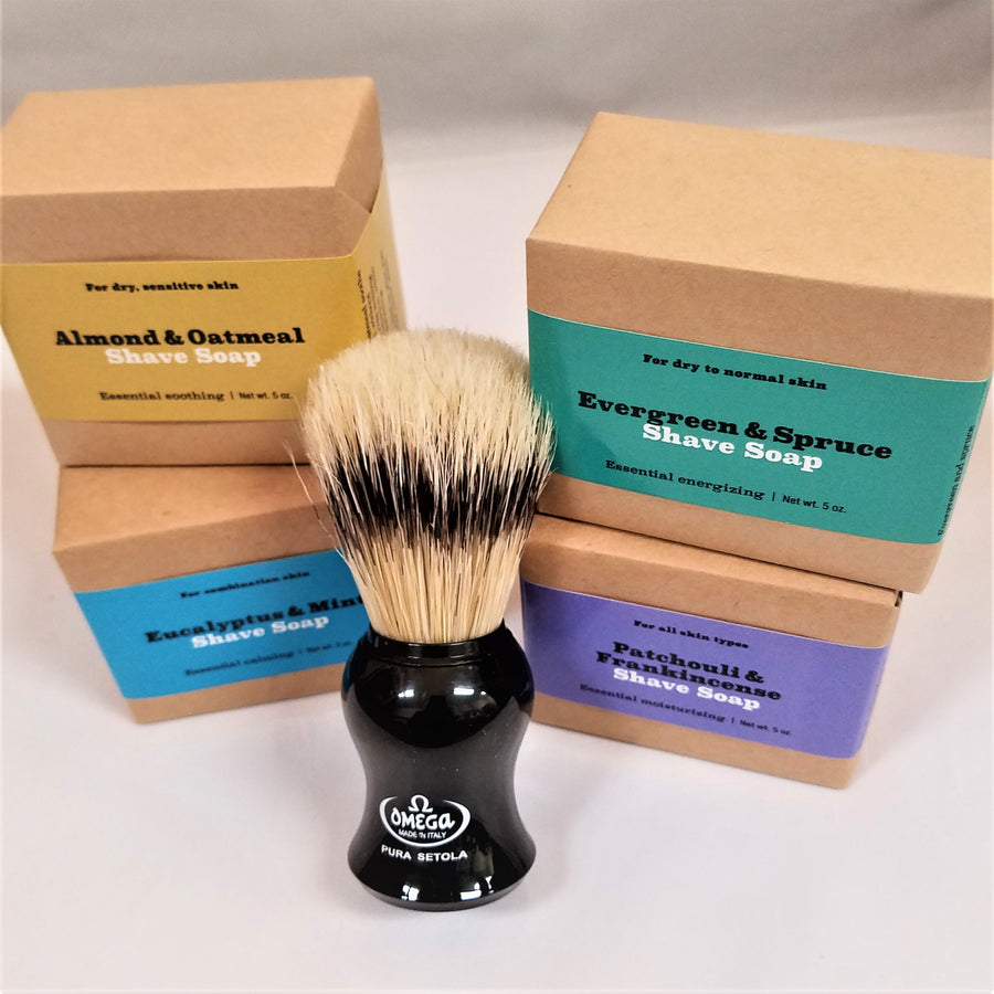 Four soaps in their wrappings piled two to the left and two to the right of a standing shaving brush with black bottom and natural and black bristles.