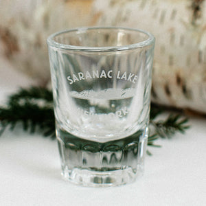 Clear shot glass with text SARANAC LAKE NEW YORK arched around mountains in the center. in front of a birch log and branch.