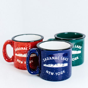 Three camp mugs red on the left, blue center, green peeking out from behind on the right. Each is speckled in design with the words SARANAC LAKE in white arched over white mountains over white lettering NEW YORK. Black rims; red and blue have white interiors; green has green interior.