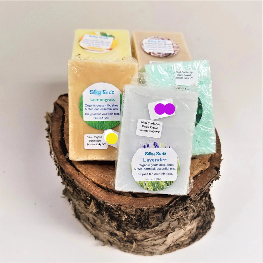 5 bars of Silky Sudz standing atop a birch tree round. Pale gray lavender-scent bar in front with Lemongrass bar to the left. Pale green bar behind the lavender and a yellow bar and light beige bar behind that.