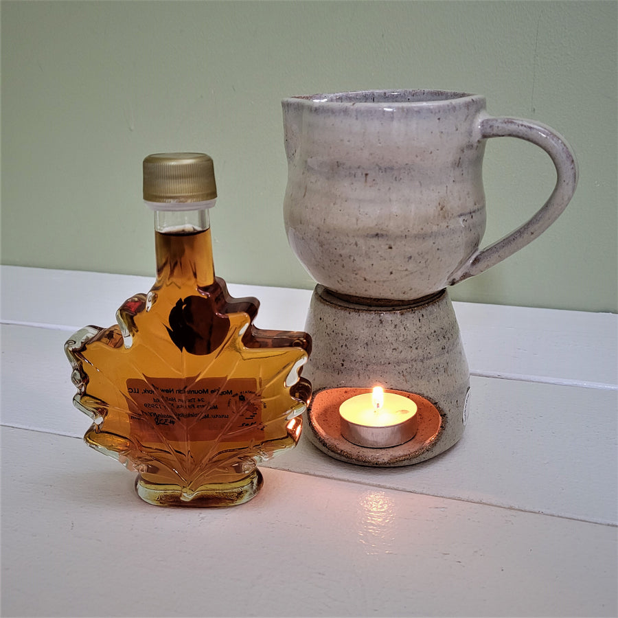 Glass maple leaf syrup bottle with a gold twist top stands to the left of a two-piece maple syrup warmer. The bottom of the warmer holds a lit tea light seen through an oval opening in the ceramic bottom. Sitting atop this is a mini ceramic pitcher with a handle on the right side.