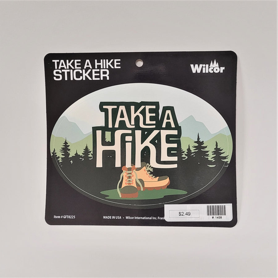 Oval TAKE A HIKE sticker on its card pacaking. The type is white with a green outline centered in a background of pale pastel mountains and dark green evergreens. A pair of beige hiking boots sits center under the type on top of a green puddle.