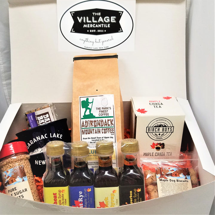 White box open to show Village Mercantile logo onto and an assortment of products in the body of the box: red-capped pure maple sugar nuggets, four barrel-aged syrups standing together, plastic package of Maple Dog Biscuits. Behind that a white box of Birch Boys Maple Chaga Tea, next to a beige package of Adirondack Mountain Coffee, next to a black Saranac Lake mug with a Dak Bar sticking out ot it.