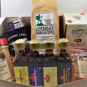 White box open to show Village MClose up of the product in the body of the box: red-capped pure maple sugar nuggets, four barrel-aged syrups standing together, plastic package of Maple Dog Biscuits. Behind that a white box of Birch Boys Maple Chaga Tea, next to a beige package of Adirondack Mountain Coffee, next to a black Saranac Lake mug with a Dak Bar sticking out ot it.