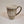Tall tan mug with whie stripe around the top with white imprint featuring the letters ADK with the A in the style of a pine tree and a small hiker climbing the top of the K. Extra large handle visible on the right side.