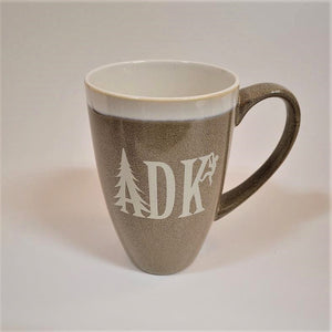 Tall tan mug with whie stripe around the top with white imprint featuring the letters ADK with the A in the style of a pine tree and a small hiker climbing the top of the K. Extra large handle visible on the right side.