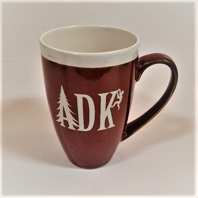 Tall burnt orange mug with whie stripe around the top with white imprint featuring the letters ADK with the A in the style of a pine tree and a small hiker climbing the top of the K. Extra large handle visible on the right side.