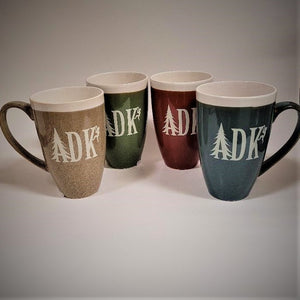 Four ADK terra bella mugs standing. From left to right tan with white imprint on front and wide handle pointing to the left side; olive green with most of imprint showing on the mug, no handle showing; burnt orange with most of white imprint showing, no handle showing; slate blue mug with white imprint showing and large handle extending to the right.