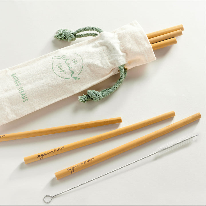 Cloth back with pale green lettering and drawstring rope with 3 bamboo straws popping out from inside. Next to the bag, lying flat are 3 bamboo straws and a silver straw cleaner with bristles on the end.
