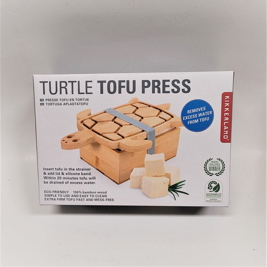 Cover of the Turtle Tofu Press box. Type is gray on white with a blue circle with white type: Removes excess water from tofu and a red vertical band on the side with white type: KIKKERLAND.  A photo of the product is in the middle of the box cover with four tofu cubes placed in front of it.