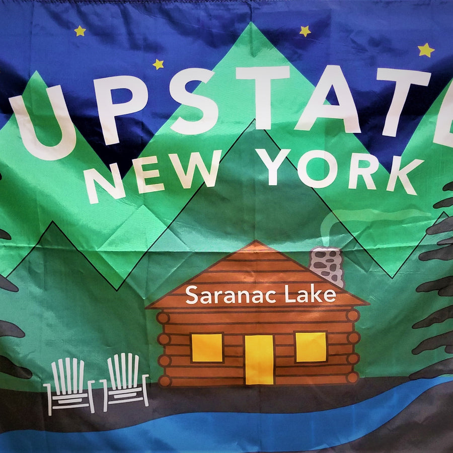 Colorful fabric with very graphic image of log cabin with Saranac Lake printed in white on the cabin. Two white Adirondack chairs next to the cabin. Green triangle graphics behind the cabin. Black and blue below; blue above with four gold stars and the works UPSTATE NEW YORK in two lines in white type over the cabin.