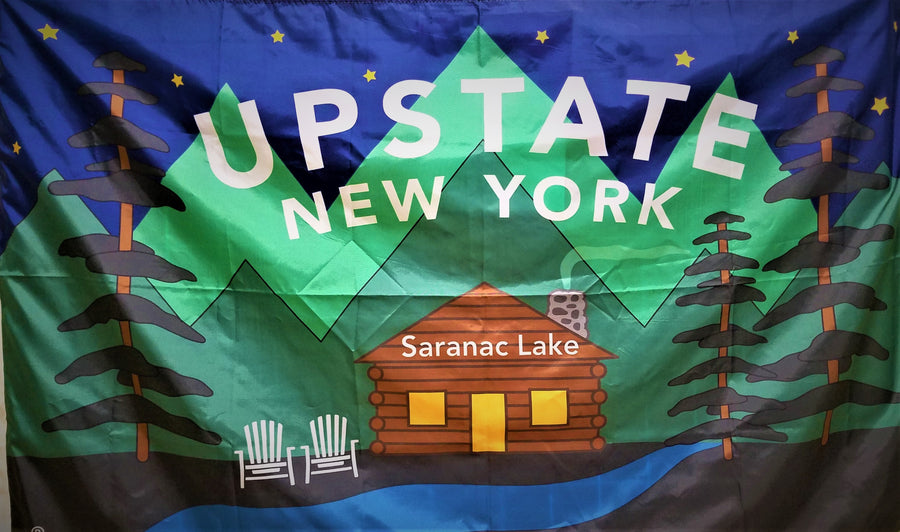 Full rectangular view of the UPSTATE NEW YORK Saranac Lake Flag. Blue top with nine gold stars showing above green triangular graphics with 3 evergreen trees surrounding the log cabin, two white Adirondack chairs and white type saying UPSTATE NEW YORK. All above a blue ribbon.